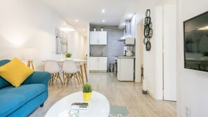 A modern living room with a blue sofa, a white coffee table, and a dining area with a small table and chairs. A kitchen with white cabinets and a stainless steel range hood is visible in the background. - G33 (Barcelona)