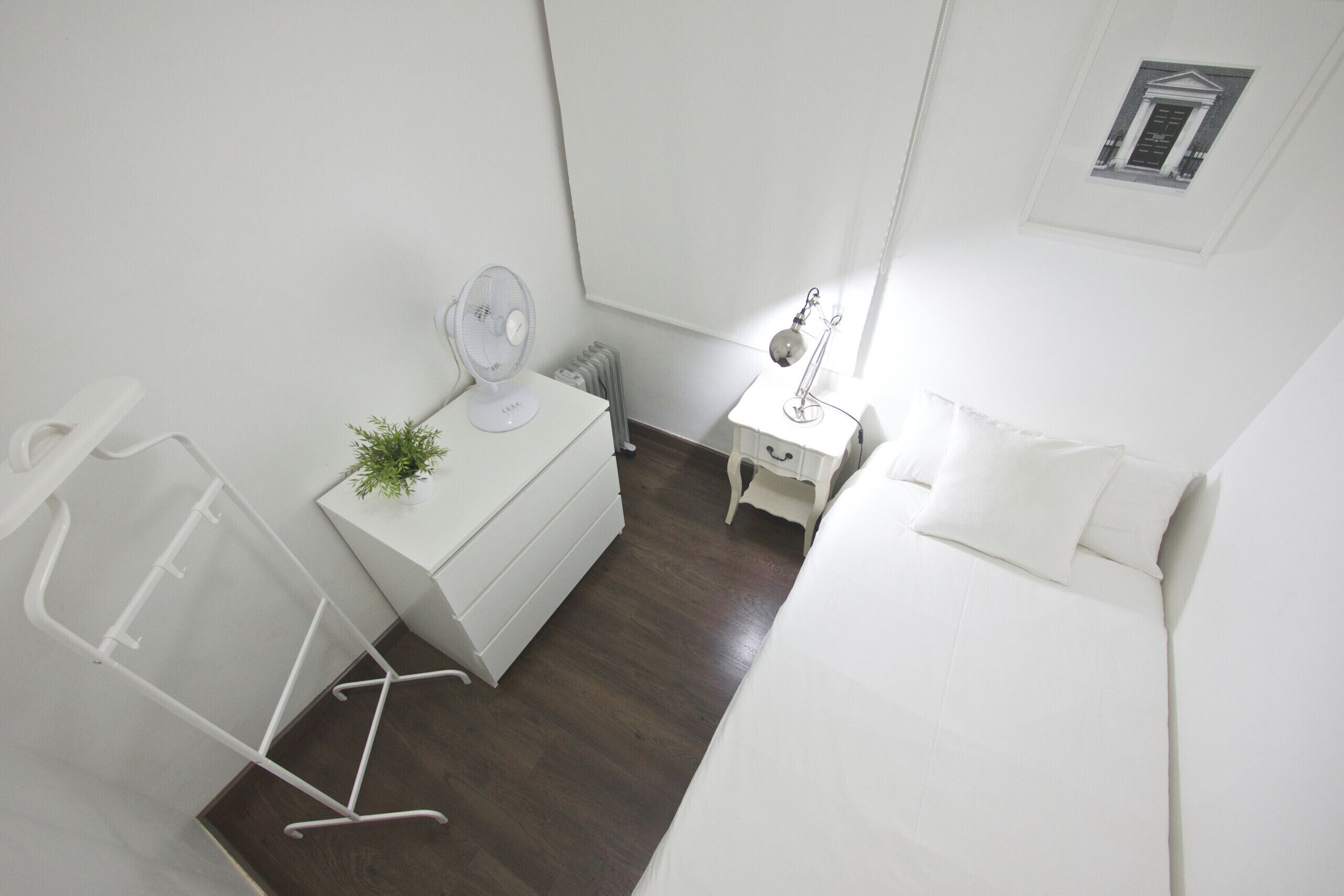 Une petite pièce blanche, avec un lit, une table de chevet dotée d’une lampe, une commode et un radiateur.