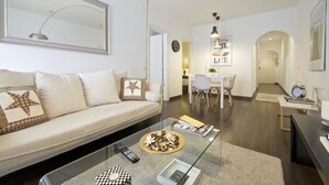 A modern living room with a glass coffee table, a sofa with decorative pillows, a dining area with a table and chairs, and a hallway leading to another room.