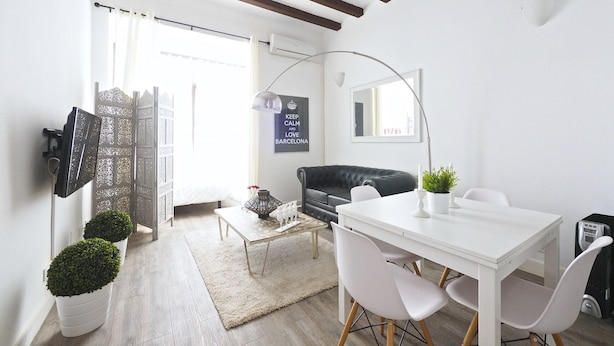 Ein modernes Wohnzimmer mit einem weißen Sofa, einem Esstisch mit Stühlen, einem Couchtisch, einem an der Wand montierten Fernseher und einer Topfpflanze.