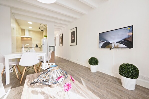 Una cocina moderna con muebles blancos, una zona de comedor con sillas blancas, un televisor de pantalla plana grande montado en la pared y una escultura decorativa de metal en el suelo.