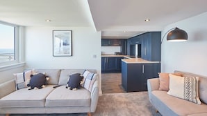 A modern living room with a sofa and a couch, a kitchen with blue cabinets, and a large window. - Host & Stay | Penthouse Pier View (Whitby)