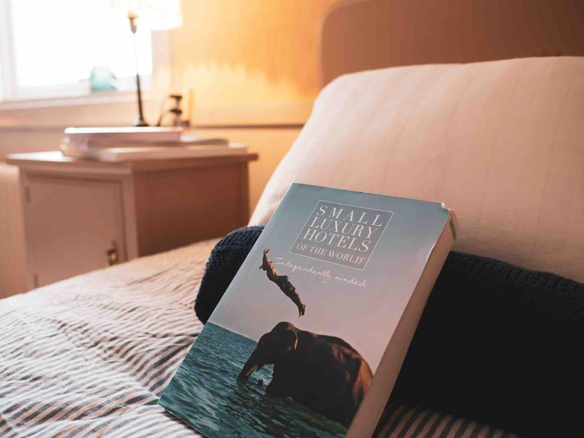 A book titled "Small Luxury Hotels of the World" on a bed with striped bedding, a nightstand, and a lamp in a hotel room.
