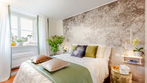 A neatly arranged bedroom with a bed, bedside tables, a potted plant, and a shelf with books and a tray of pastries.
