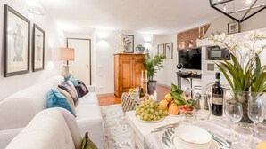 A well-lit living room with a sofa, a dining table set with fruits and glasses, a TV, and a kitchen area with a microwave.