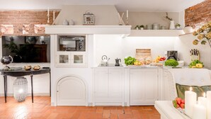 A compact kitchen with white cabinets, a microwave, and a kettle. There is a small dining area with a table and chairs, and a brick wall in the background.