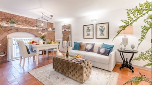 A living room with a white sofa, a tufted ottoman, a dining table with chairs, a brick wall, and a fireplace.