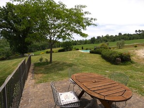 A patio with a wooden table and metal chairs, surrounded by a brick wall and a metal fence, overlooking a green landscape with a pool.