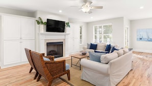 A spacious living room with a fireplace, a ceiling fan, and a large TV mounted above it. - Captain Champs Castaway | Elevator & Game Room! (Tybee Island)