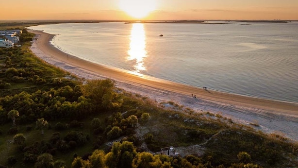 A coastal landscape with a beach, a small boat, and a few people walking along the shore. - Captain Champs Castaway | Elevator & Game Room! (Tybee Island)