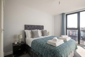 Ein modernes Schlafzimmer mit einer gesteppten Kopfteile, einem großen Fenster mit Blick auf die Stadt, einem ordentlich bezogenen Bett mit einer grünen Decke und einem Nachttisch mit einer Lampe und einem kleinen Dekorationsgegenstand.
