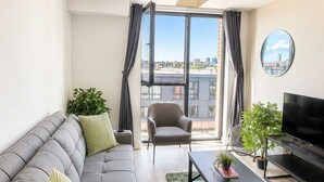 En moderne stue med sofa, lænestol, sofabord, fjernsyn og store vinduer med udsigt over byen.