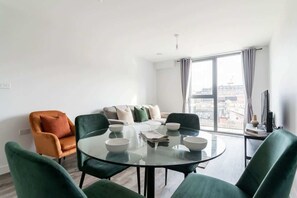 Una sala moderna con una mesa de centro de vidrio, sillas verdes, un sofá con cojines y vistas a través de amplias ventanas.
