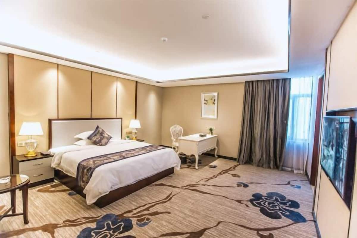 Habitación de hotel con cama, mesita de noche, lámpara, silla y un ventanal con cortinas.