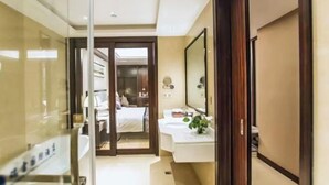 A hotel room with a glass partition, a bed, a bathroom with a sink and mirror, and a wooden door.