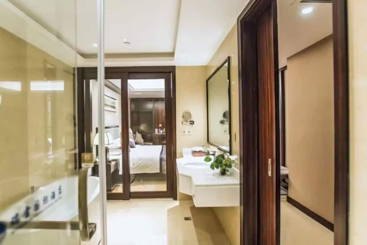 Habitación de hotel con un separador de vidrio, una cama, un baño con lavamanos y espejo, y una puerta de madera.
