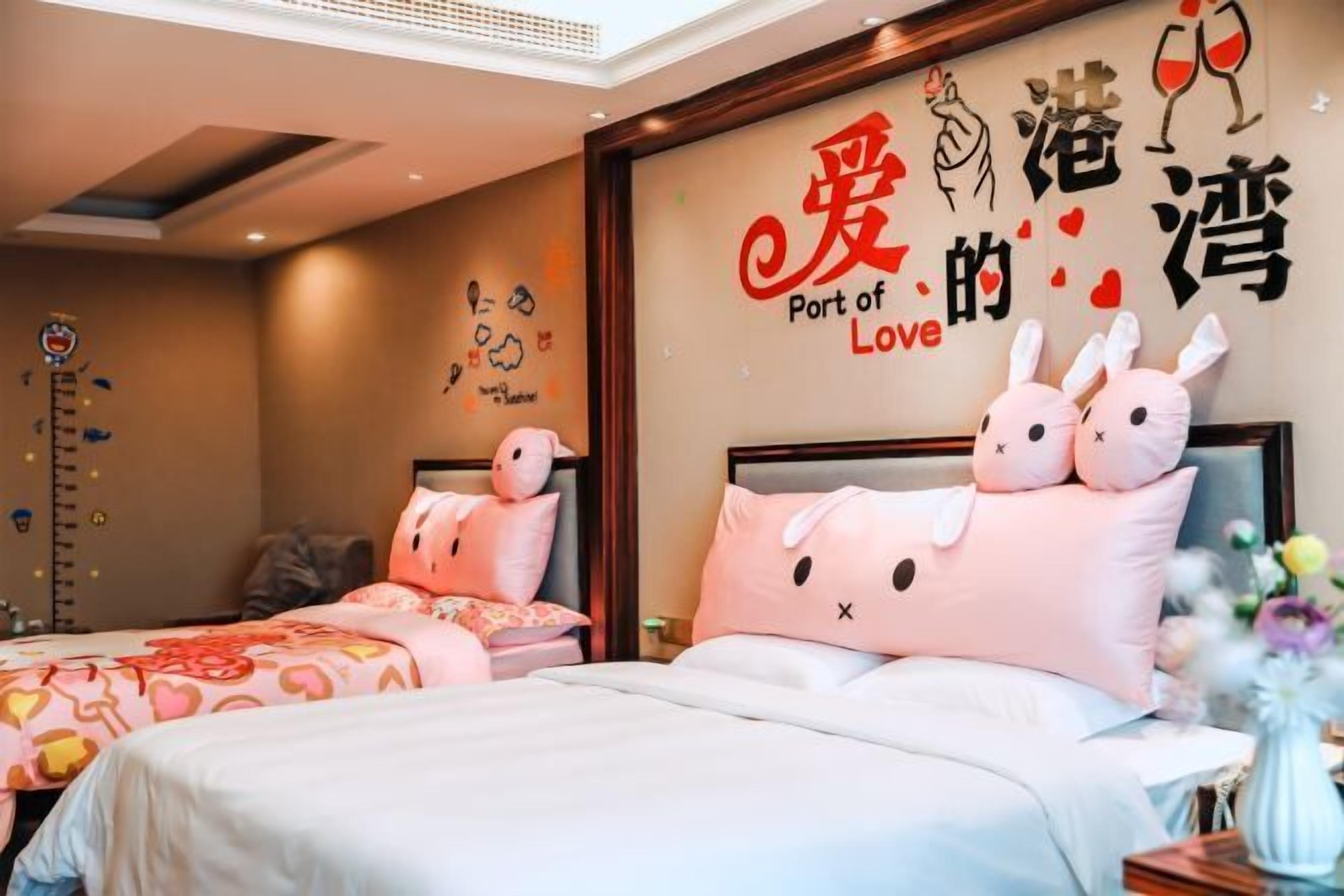 A hotel room with two beds, each with pink bedding and a bunny-themed pillow. A wall decoration reads "Port of Love" with illustrations of hearts and wine glasses.