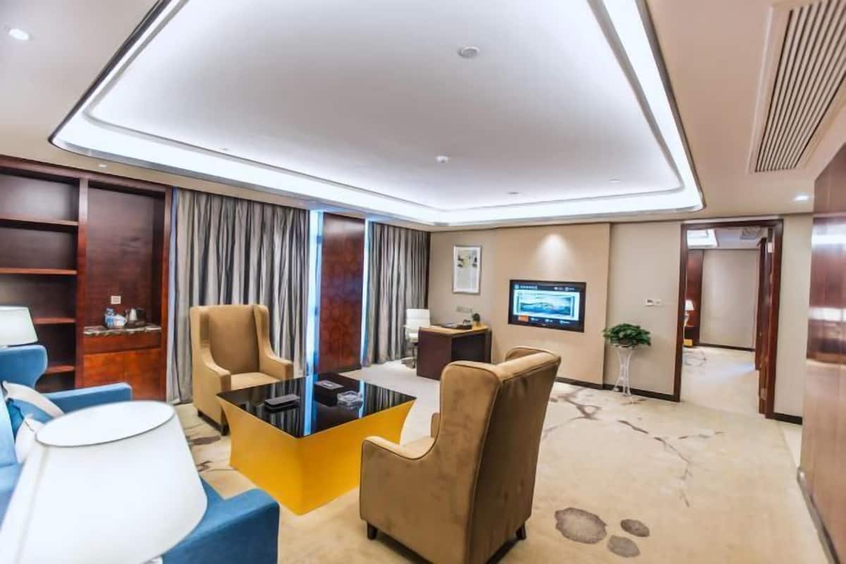 Habitación de hotel moderna con sofá, sillones, una mesa de centro, televisor y un ventanal grande con cortinas.