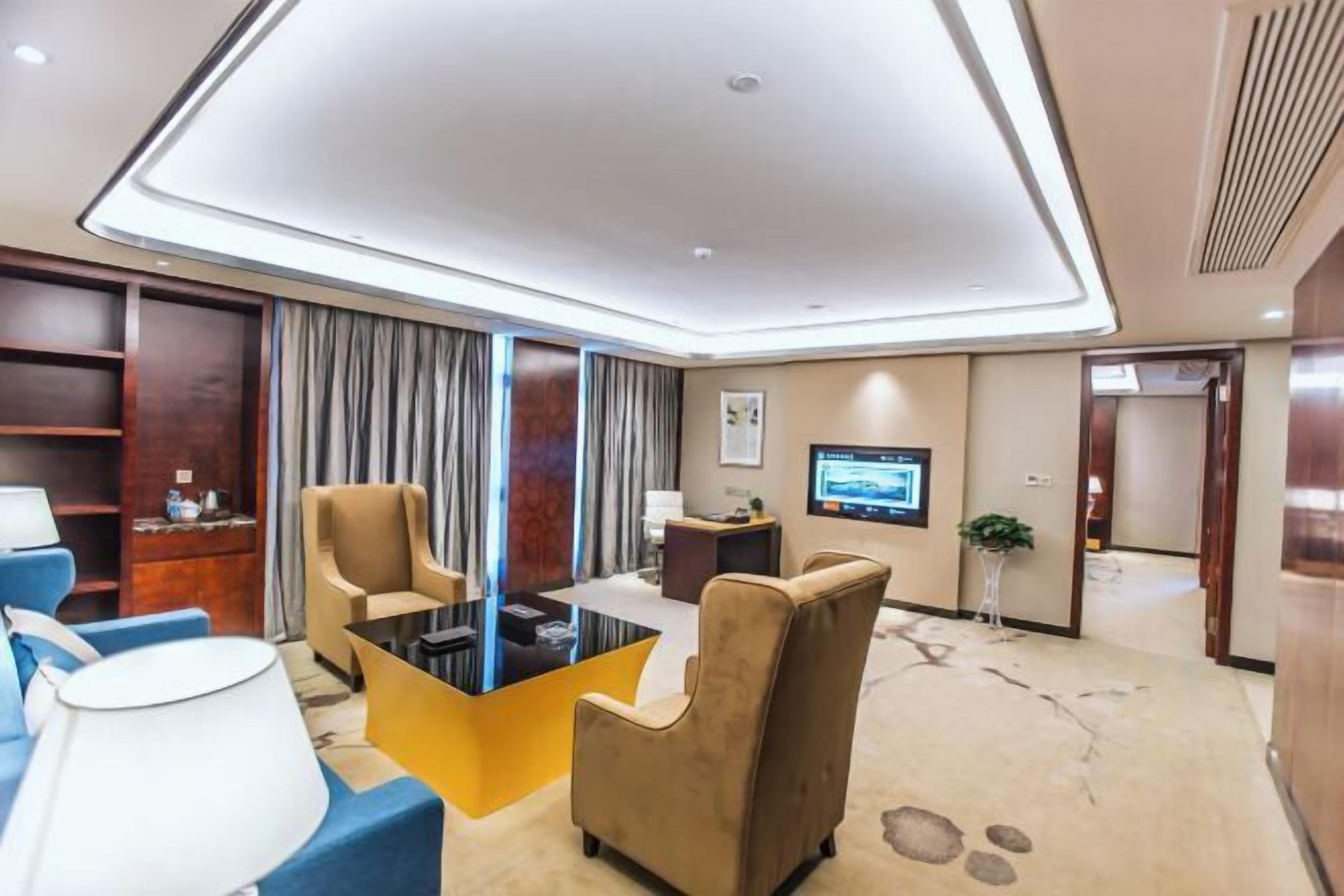 A modern hotel room with a large flat-screen TV, comfortable seating, a coffee table, and a large window with curtains.