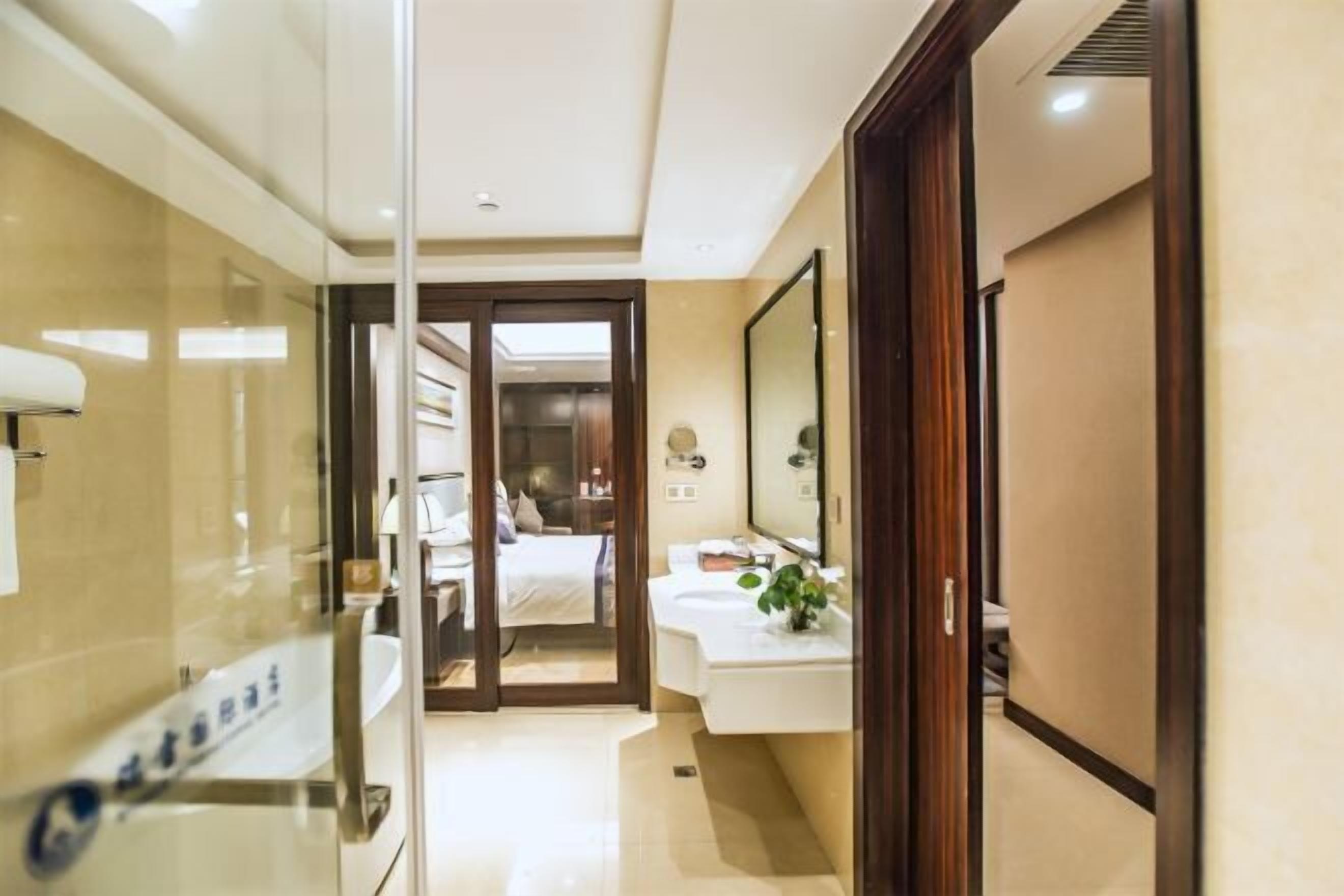 A hotel bathroom with a glass partition, a sink, a mirror, and a bed visible through an open door.