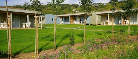A row of modern, single-story buildings with attached outdoor seating areas, surrounded by well-maintained lawns and a variety of flowering plants.