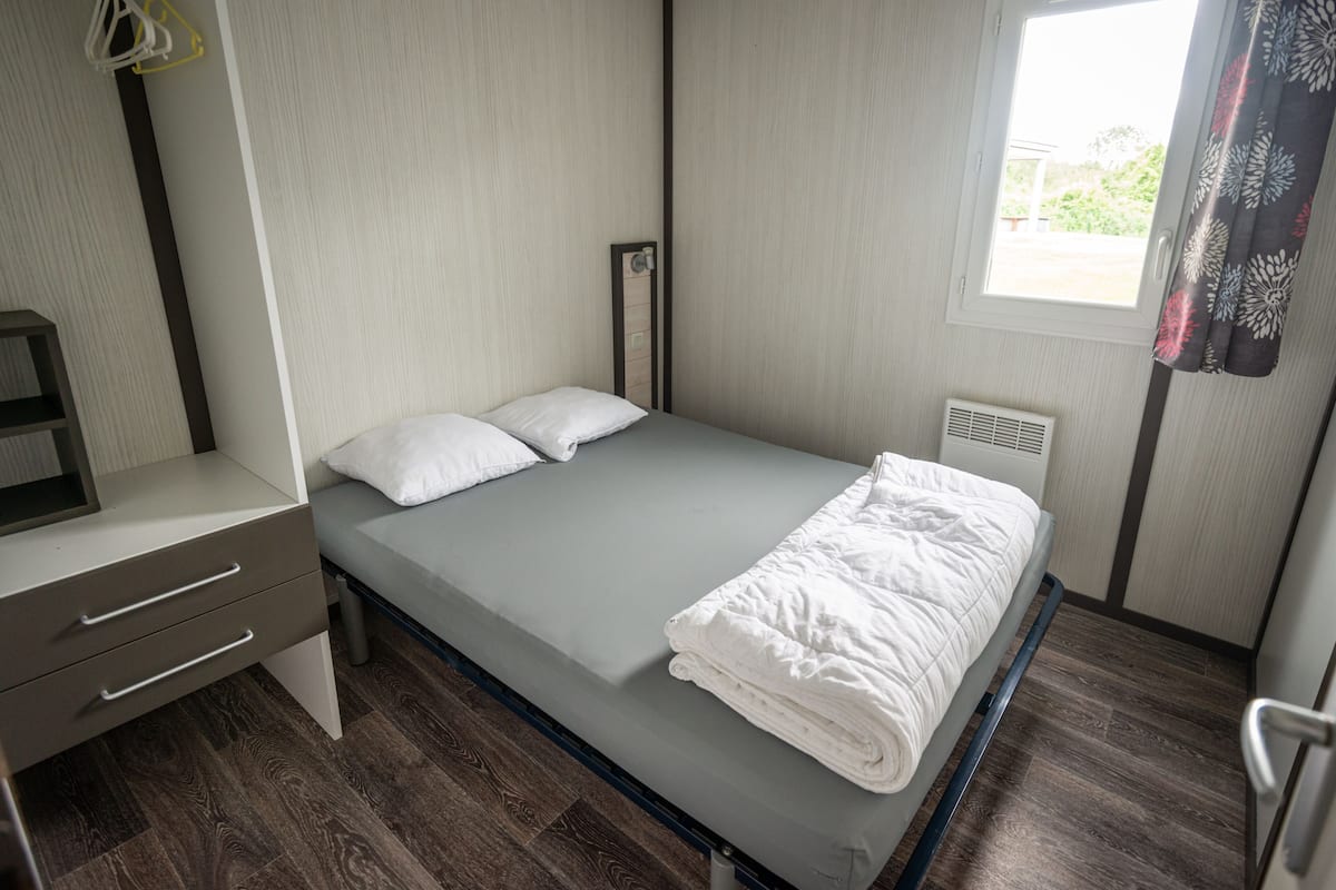 A single bed with a white mattress and pillows, a bedside table with drawers, a window with curtains, and a radiator.