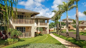 A two-story residential building with a balcony, surrounded by well-maintained lawns and palm trees.