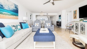 A living room with a sofa, ottoman, and chairs, a ceiling fan, and a TV mounted on the wall. - Stay @ Our Folly Place on Folly Beach (Folly Beach)