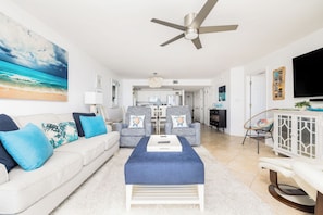 A living room with a sofa, ottoman, and chairs, a ceiling fan, and a TV mounted on the wall.