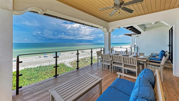 A covered outdoor area with a wooden ceiling, a ceiling fan, a table with chairs, and a sofa. It overlooks a beach with the ocean and a clear sky.