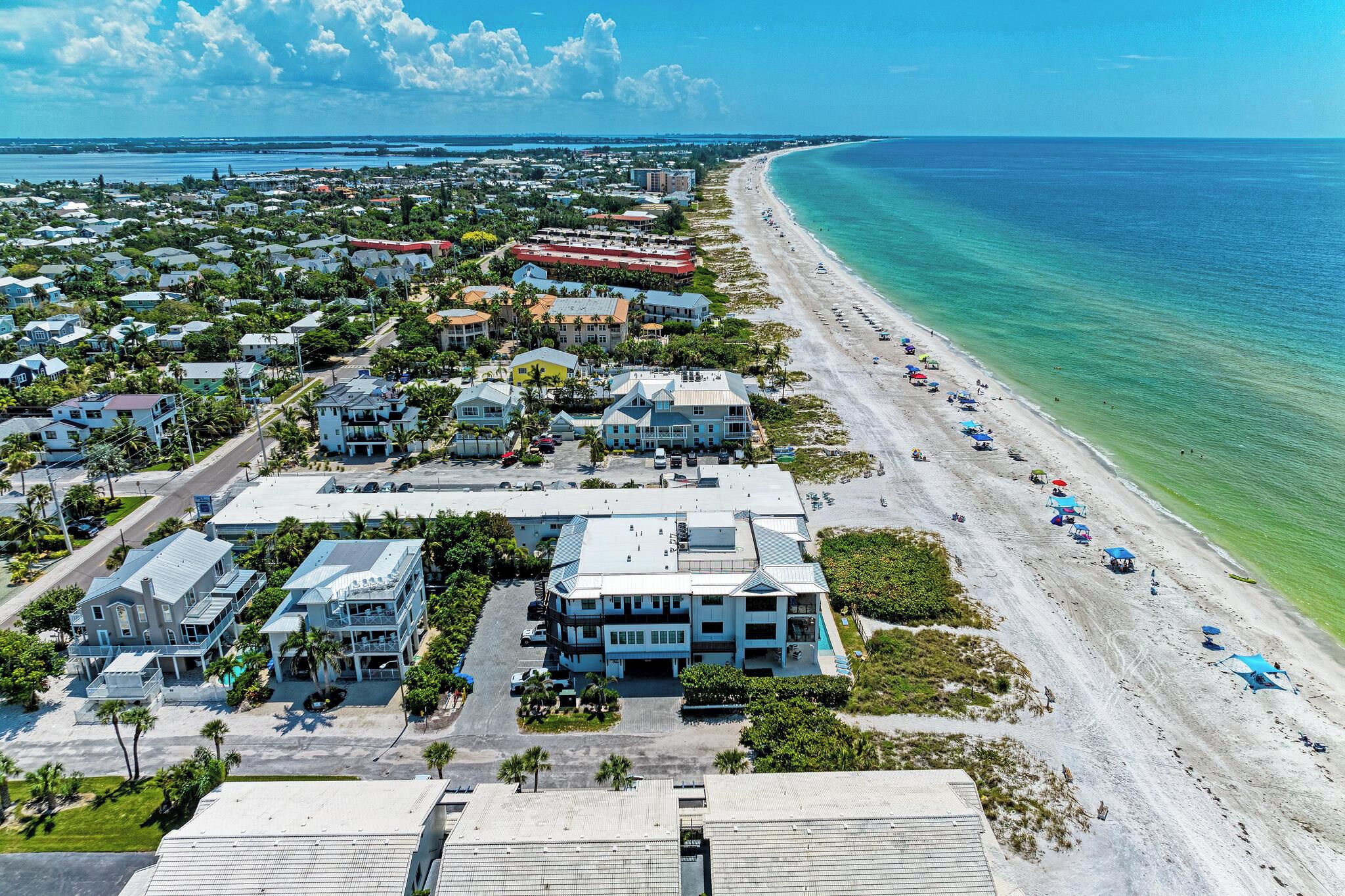 An aerial view of a coastal town with a beachfront, including buildings, roads, and a beach with umbrellas.