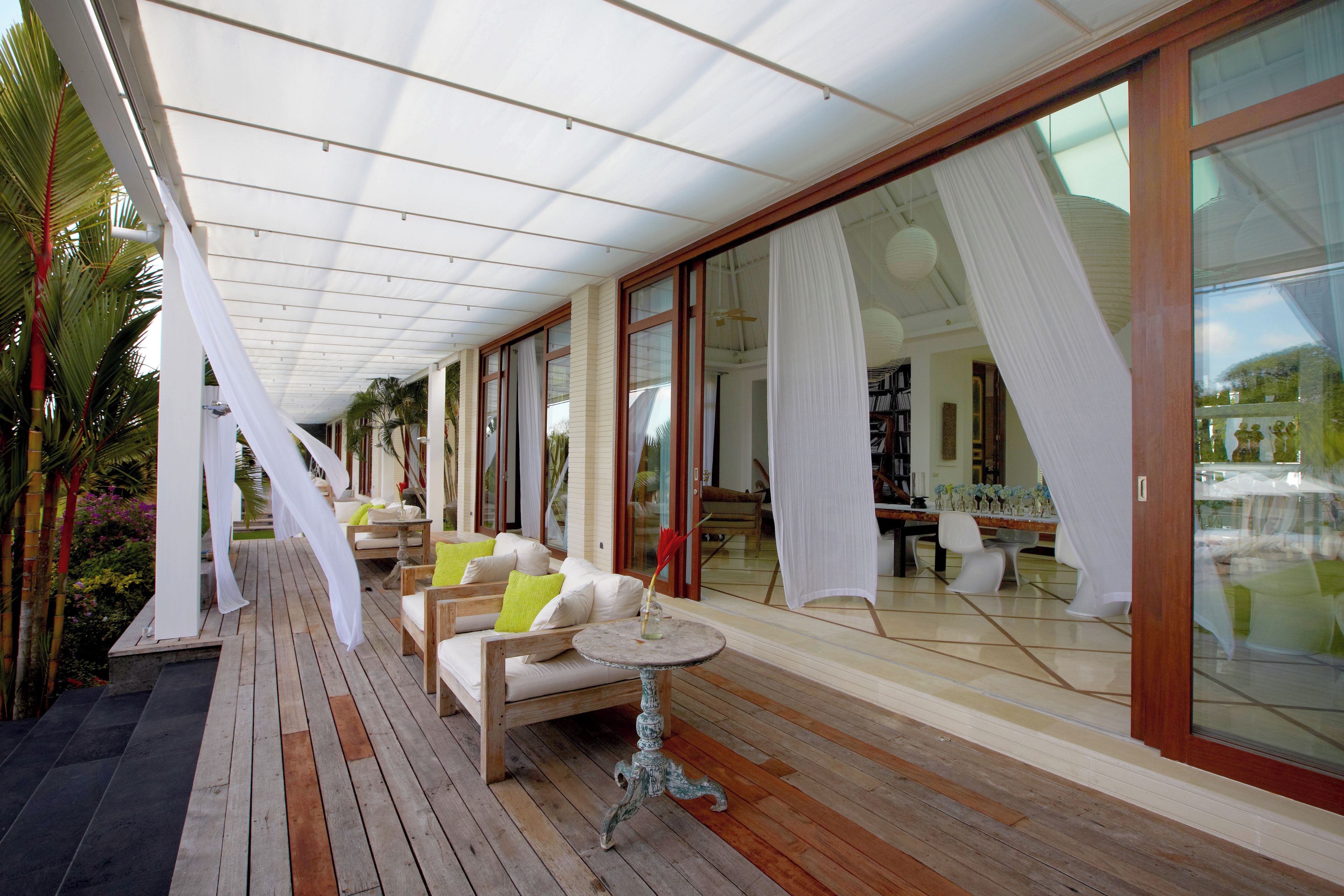 A covered outdoor seating area with wooden flooring, white curtains, and a glass wall.