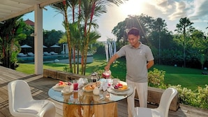 Une table en verre, entourée de chaises blanches, est dressée pour le petit-déjeuner. L’ensemble est agrémenté d’une terrasse en bois et d’une végétation luxuriante.
