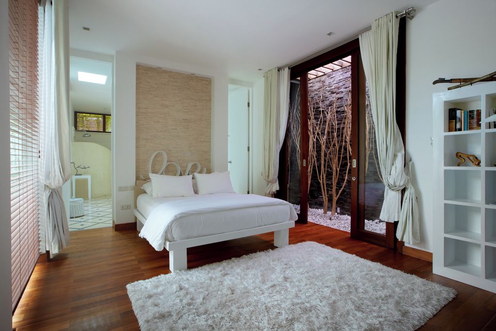 A bedroom with a white bed, wooden flooring, a white rug, a bookshelf, and a view through a glass door.