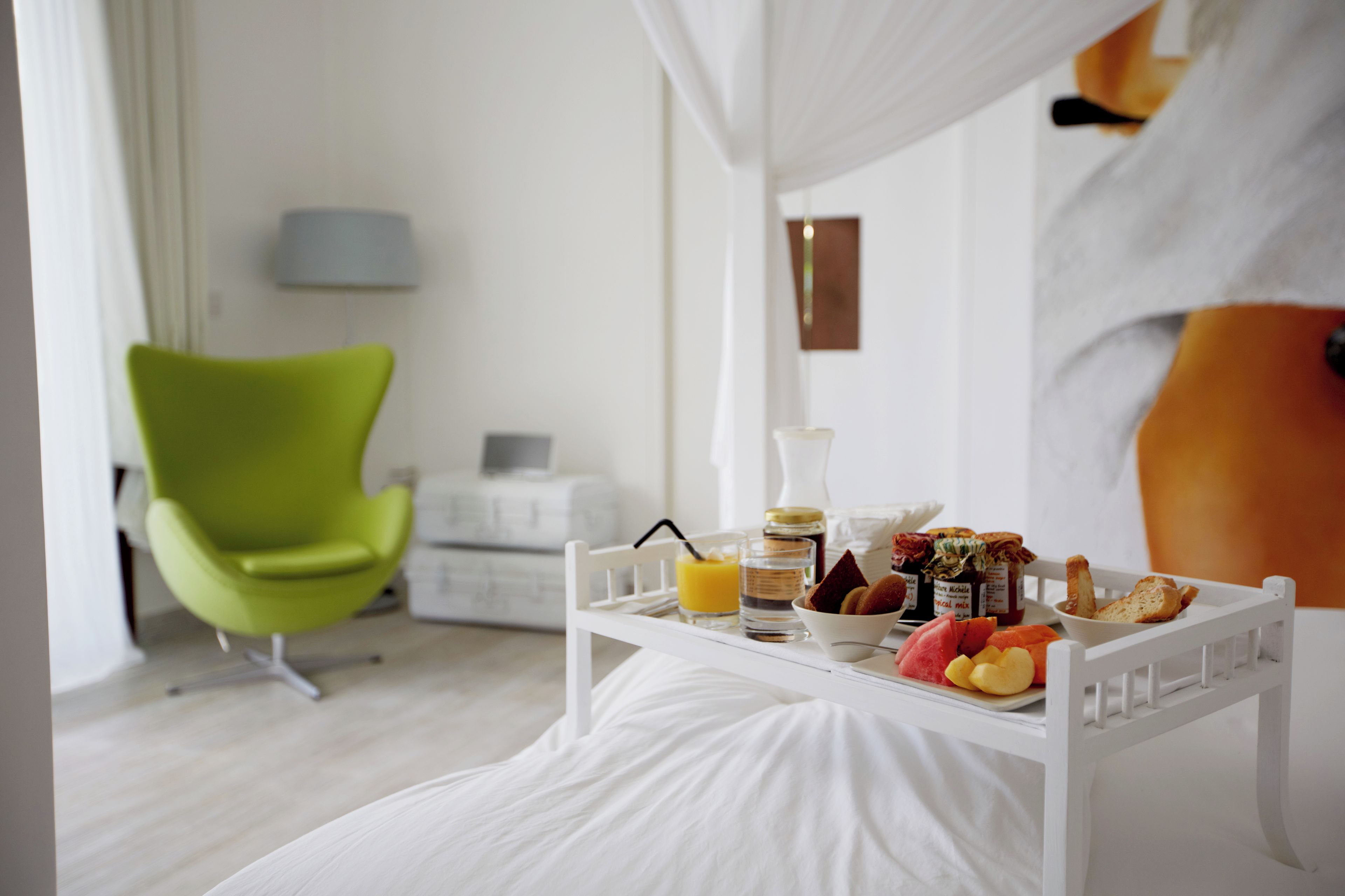 Una cama blanca con dosel, una silla verde, una mesita de noche con elementos para el desayuno y otra mesita de noche blanca con lámpara.