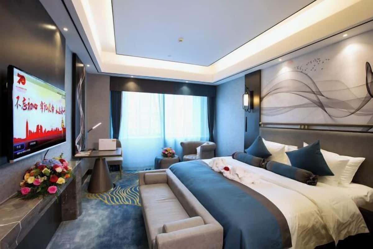 Una habitación de hotel moderna con una cama grande, un escritorio con televisor de pantalla plana, una zona de estar con sofá y sillones, y una pared decorativa con arte abstracto.