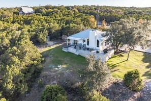 A modern house with a large deck, surrounded by dense trees and a grassy yard. - Belle Vue: Hot-Tub + 5 mins from River! (New Braunfels)