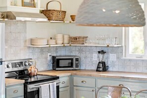 A kitchen with a stove, microwave, and kettle. There are shelves with plates and glasses, and a window above the counter. - Belle Vue: Hot-Tub + 5 mins from River! (New Braunfels)