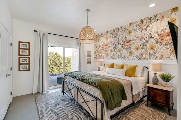 A bedroom with a bed, a large floral wallpaper, a hanging light fixture, and a view to the outside.