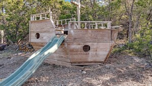 Ein Spielhäuschen in Form eines hölzernen Piratenschiffs mit Rutsche, umgeben von Bäumen.