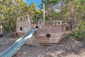 A wooden pirate ship playhouse with a slide, surrounded by trees.