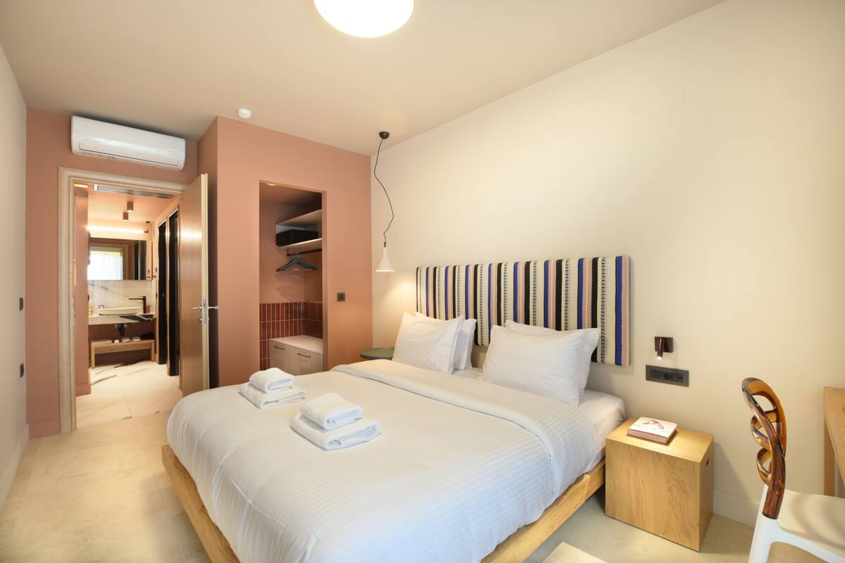 Une chambre à coucher avec un grand lit, une table de chevet, une commode, une chaise en bois, une armoire et un appareil de climatisation.