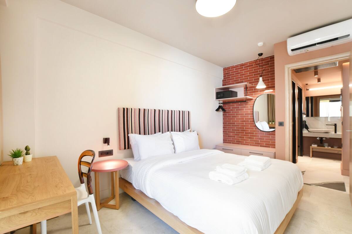 Une chambre d’hôtel avec un lit, une petite table, une chaise, un miroir et une salle de bain visible à travers une porte ouverte.