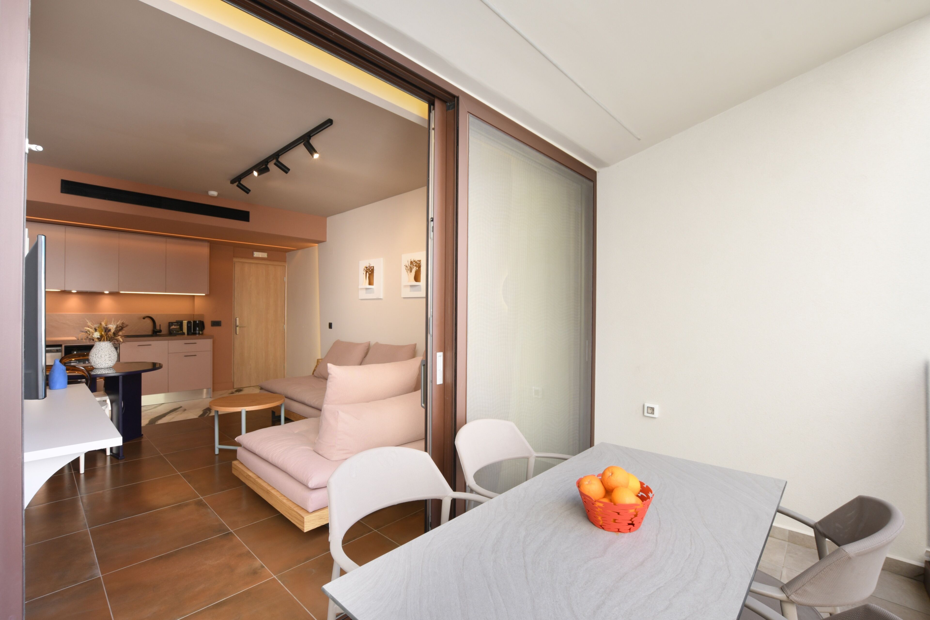 A modern living room with a sofa, a dining table with a fruit basket, and a kitchen in the background.
