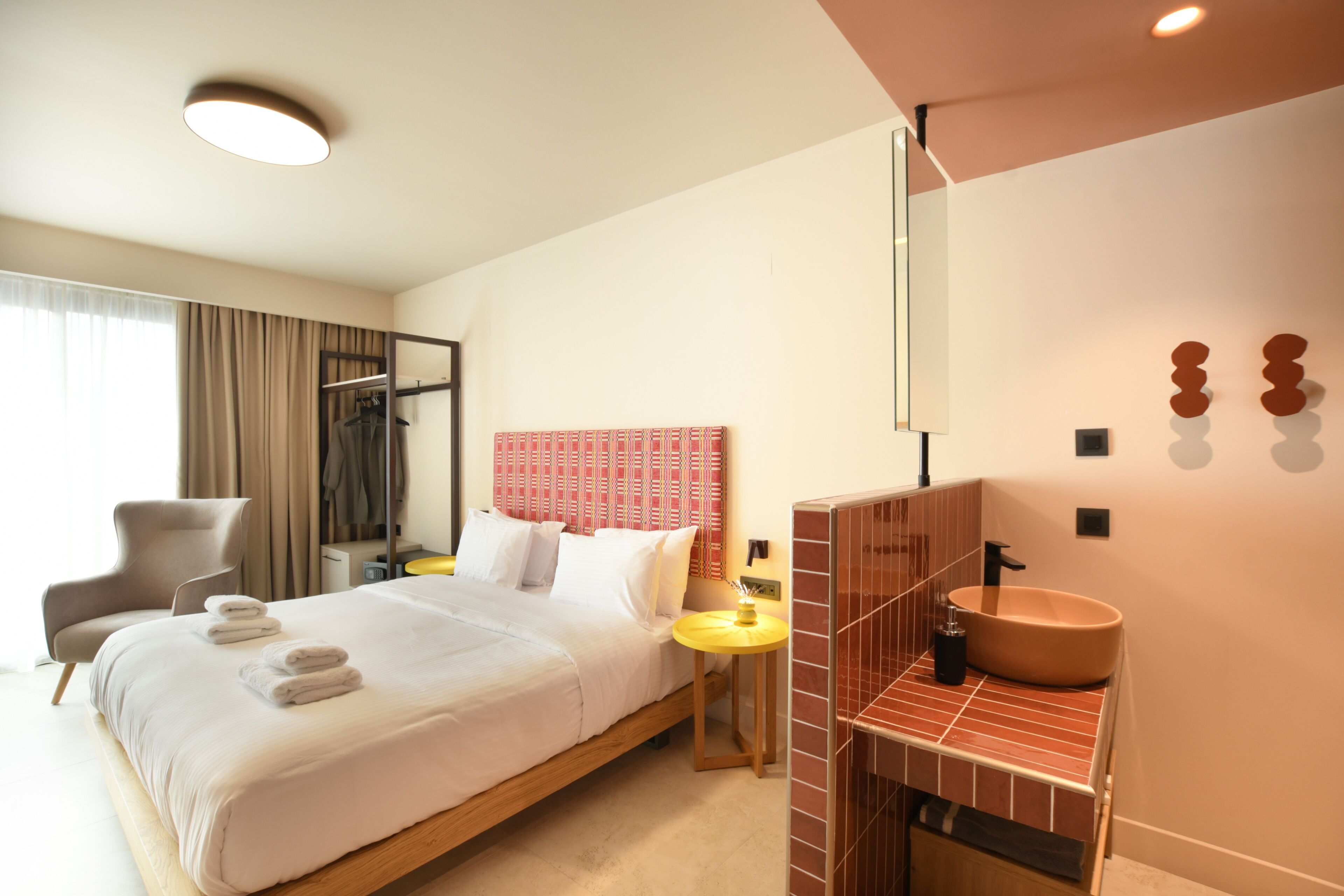 A hotel room with a bed, a chair, a bedside table, a mirror, and a bathroom with a sink and a towel rack.