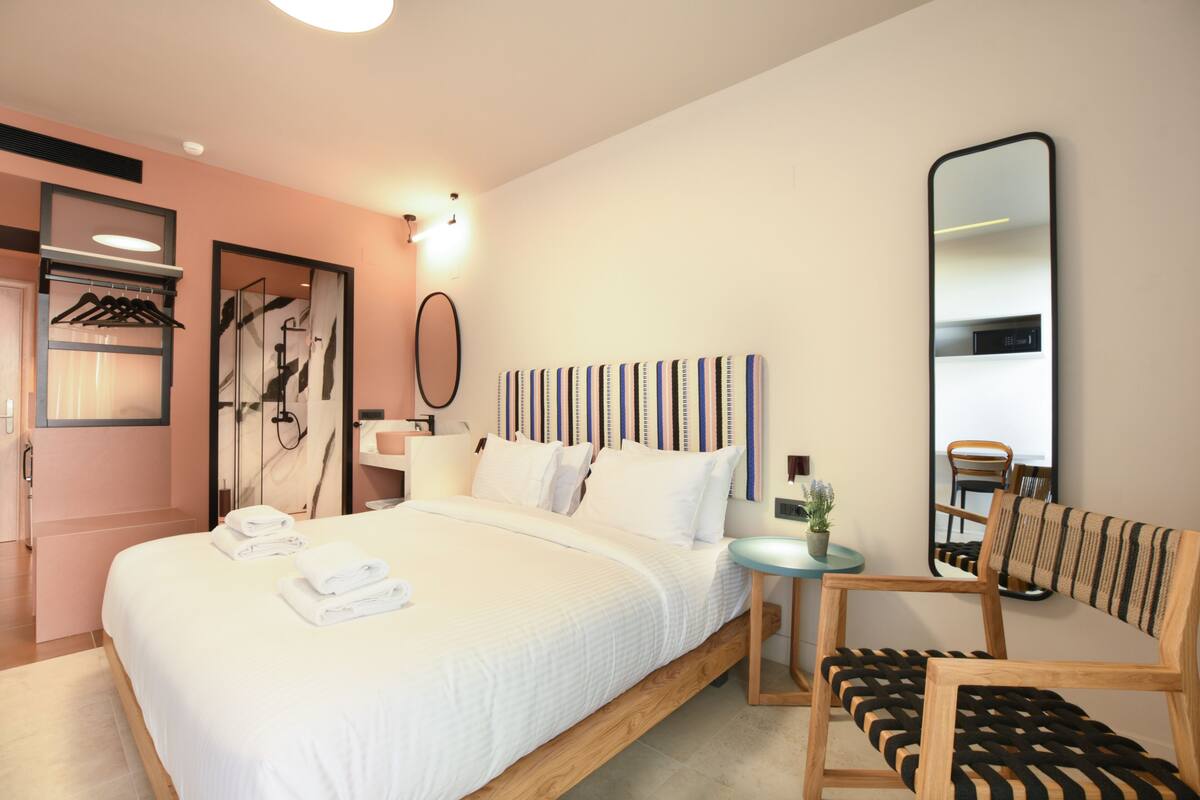 Une chambre d’hôtel moderne avec un grand lit, une chaise en bois, une petite table et un miroir au mur.