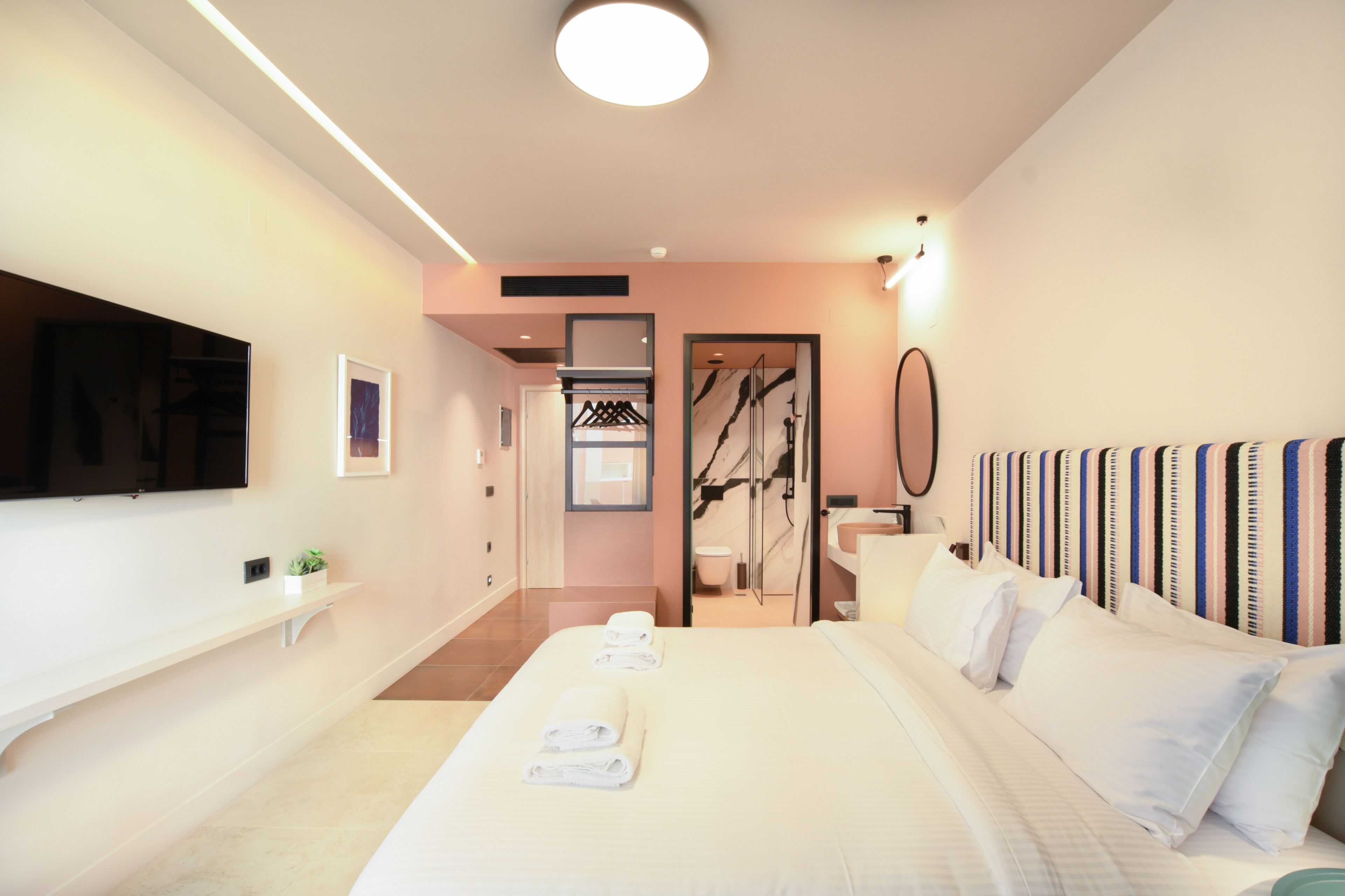 Ett modernt hotellrum med en stor säng, en platt-TV som är monterad på väggen, en liten växt på en hylla och ett badrum som syns genom en öppen dörr.