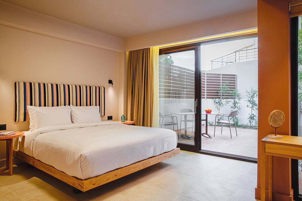Une chambre d’hôtel moderne dotée d’un grand lit, d’une tête de lit en bois, d’une petite table avec une lampe et d’une porte-fenêtre coulissante donnant sur un espace extérieur avec une table et des chaises.