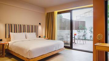 Una habitación de hotel moderna con una cama grande, cabecera de madera, una mesita con lámpara y un ventanal corredero que da a un espacio exterior con una mesa y sillas.