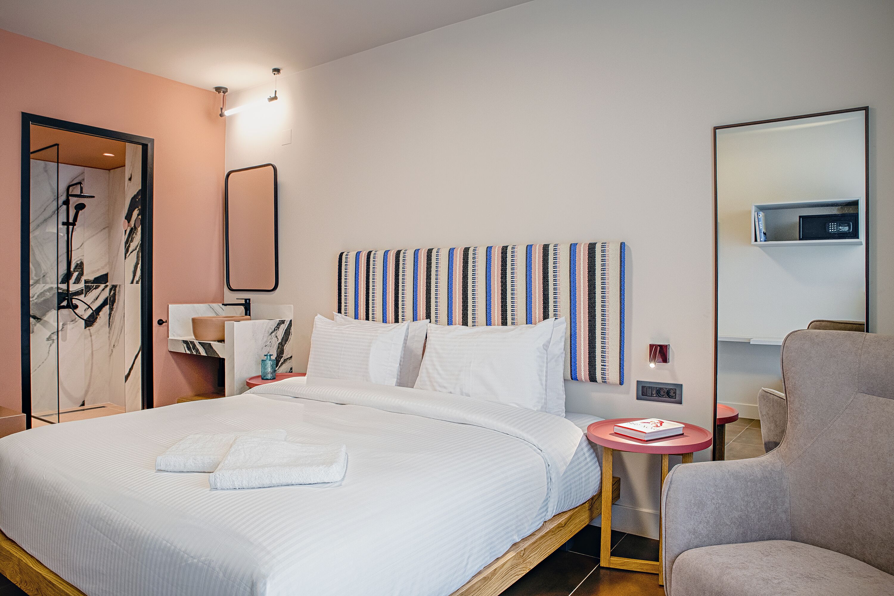 Une chambre d’hôtel moderne avec un grand lit, une table de chevet avec un livre, une chaise et une salle de bain équipée d’une douche et d’un miroir.